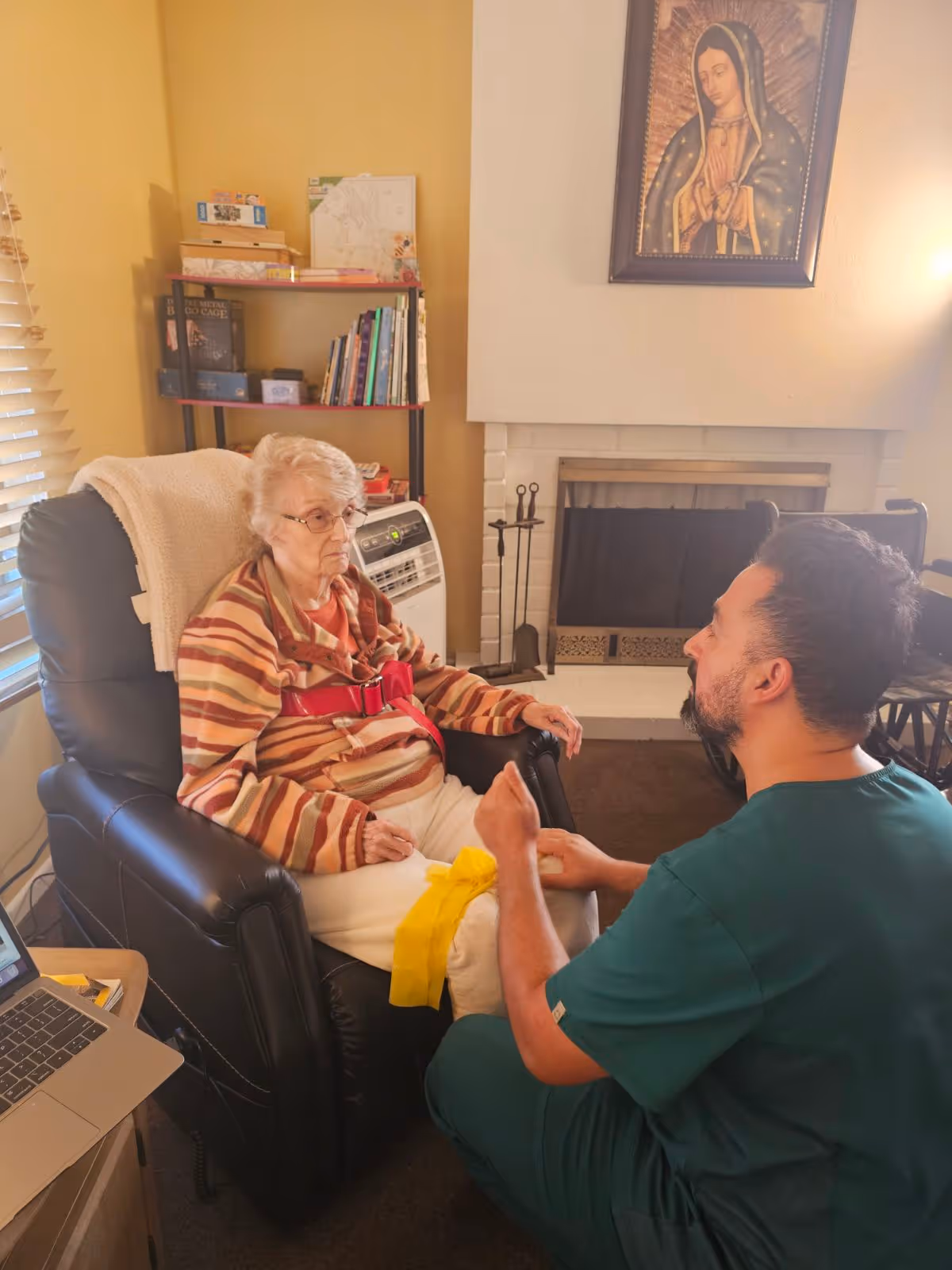 An elderly woman sitting in a black recliner chair with a striped sweater and a red belt, while a man in green scrubs kneels in front of her holding her leg, in a cozy room with a fireplace, bookshelf, and a religious painting on the wall.