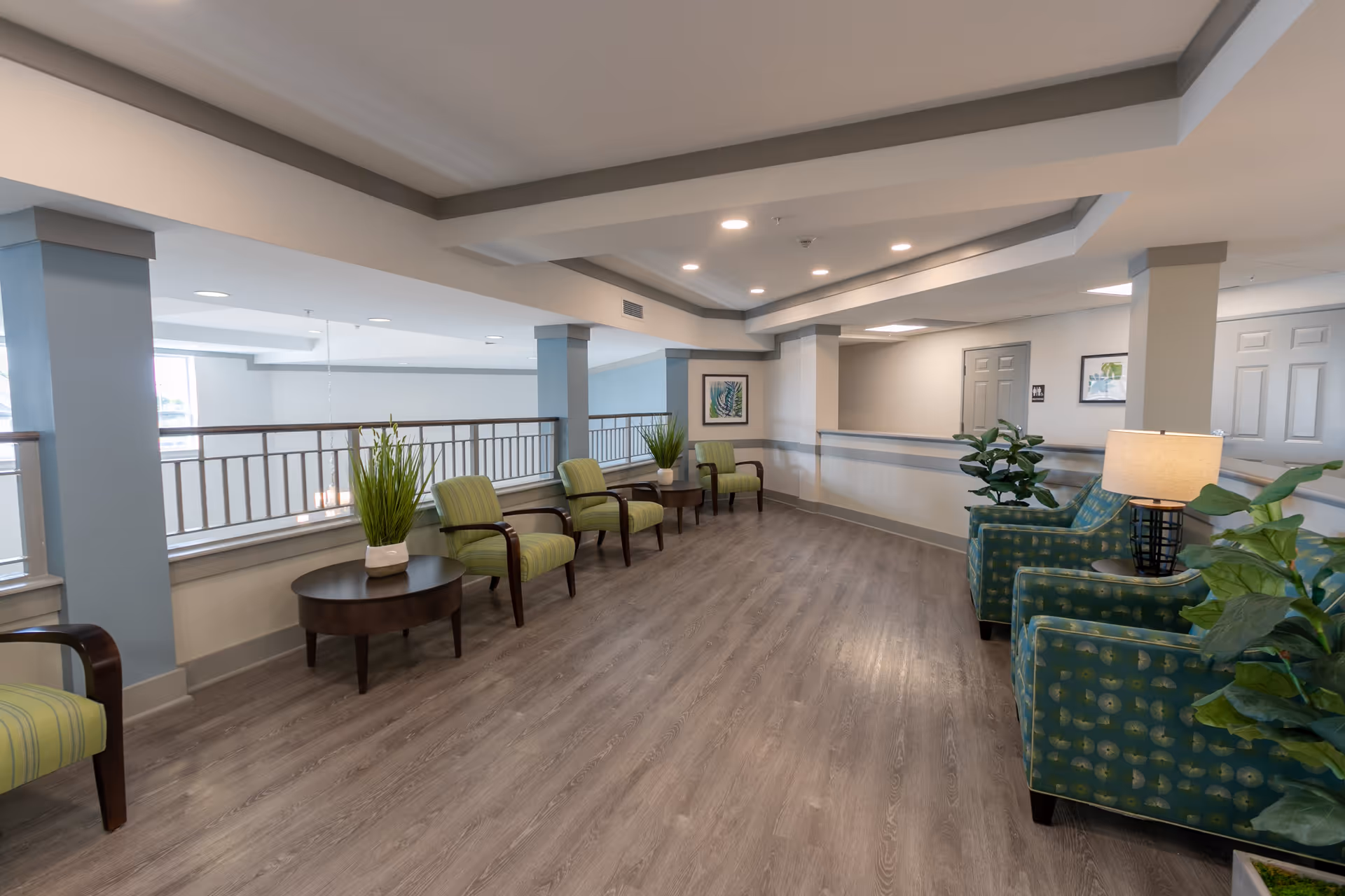 A spacious, well-lit common area with light wood flooring, green and blue upholstered chairs, small round tables with potted plants, and neutral-colored walls with framed artwork. The ceiling has recessed lighting and decorative molding.