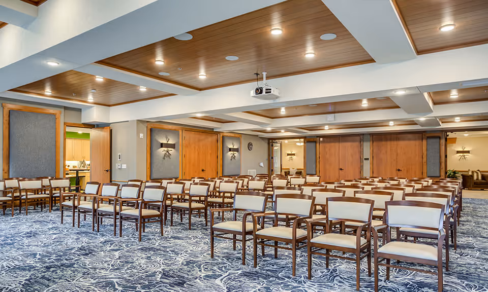 A large meeting or conference room with rows of wooden chairs with beige cushions arranged facing forward. The room has a blue patterned carpet, wooden ceiling panels with recessed lighting, and light-colored walls with wooden accents. A projector is mounted on the ceiling.