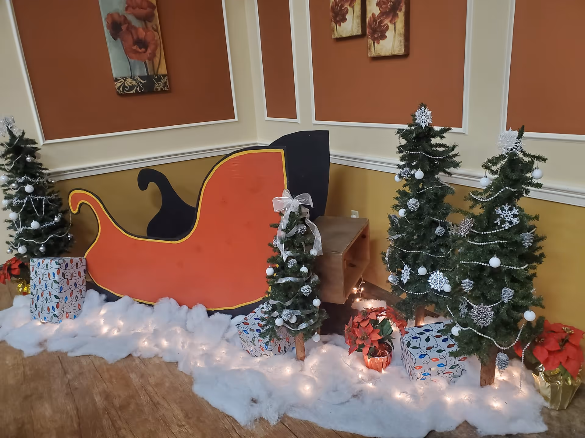 Indoor holiday display featuring a large red and black sleigh cutout surrounded by small decorated Christmas trees, wrapped gift boxes, poinsettia plants, and white artificial snow with string lights on a wooden floor against a wall with orange and cream panels and floral paintings.