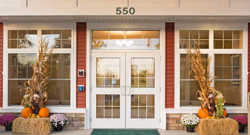 Front entrance of a building with double glass doors numbered 550, flanked by fall decorations including pumpkins, hay bales, corn stalks, and potted flowers.
