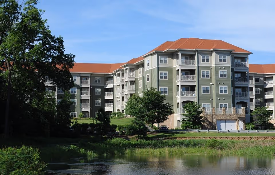 Exterior view of a multi-story senior living facility building with green and beige siding and red roofs, surrounded by trees and landscaping, with a pond in the foreground under a clear blue sky.