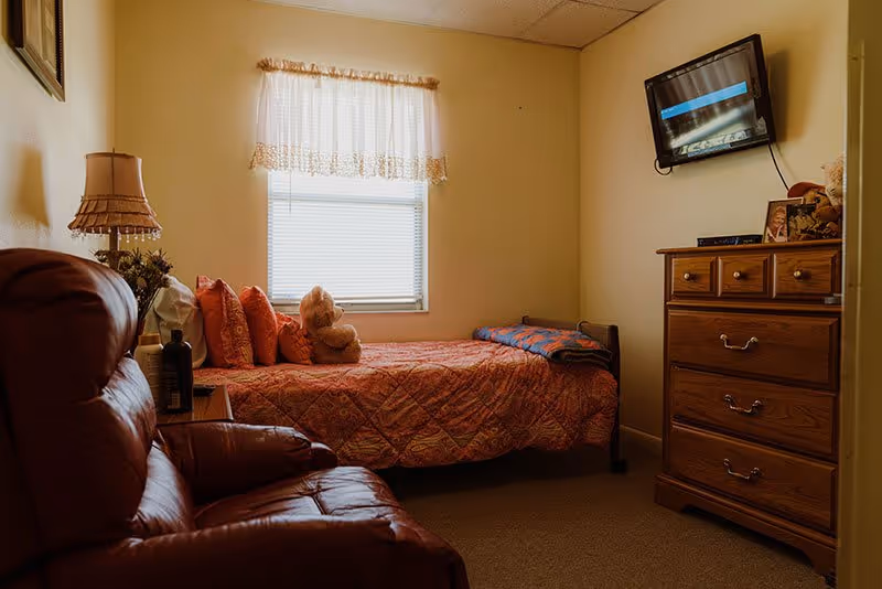 Small furnished bedroom with a single bed, teddy bear, recliner chair, wooden dresser and a wall-mounted TV.