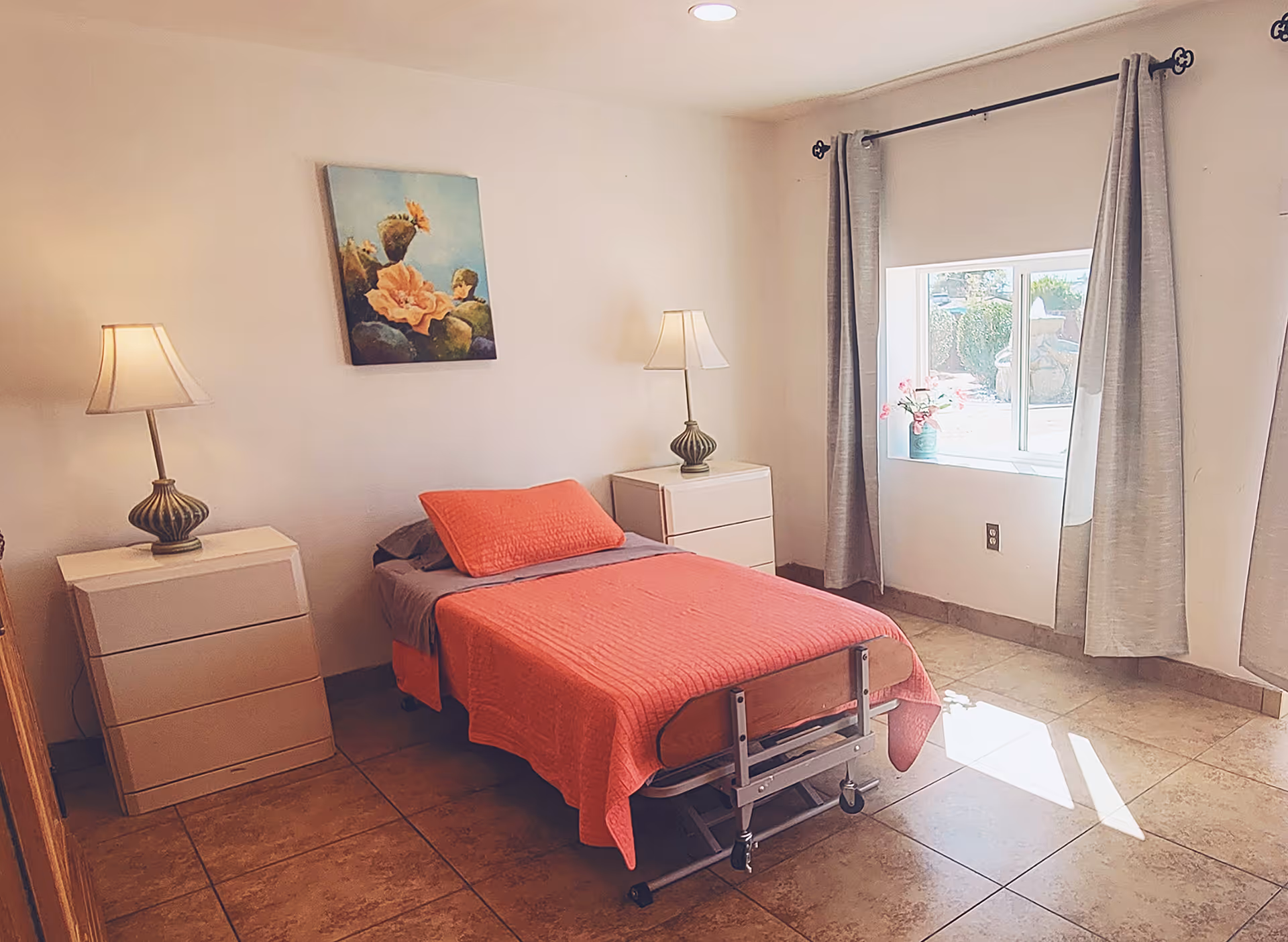 A simple bedroom with a single bed covered in a coral-colored blanket and pillow. There are two white nightstands on either side of the bed, each with a decorative lamp. A painting of flowers hangs on the wall above the bed. A window with gray curtains lets in natural light, illuminating the tiled floor.