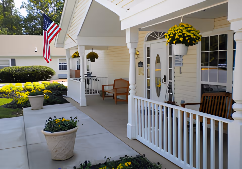 Front porch area of a building with white railings and columns, wooden chairs, hanging yellow flower pots, an American flag, and potted plants along the walkway.