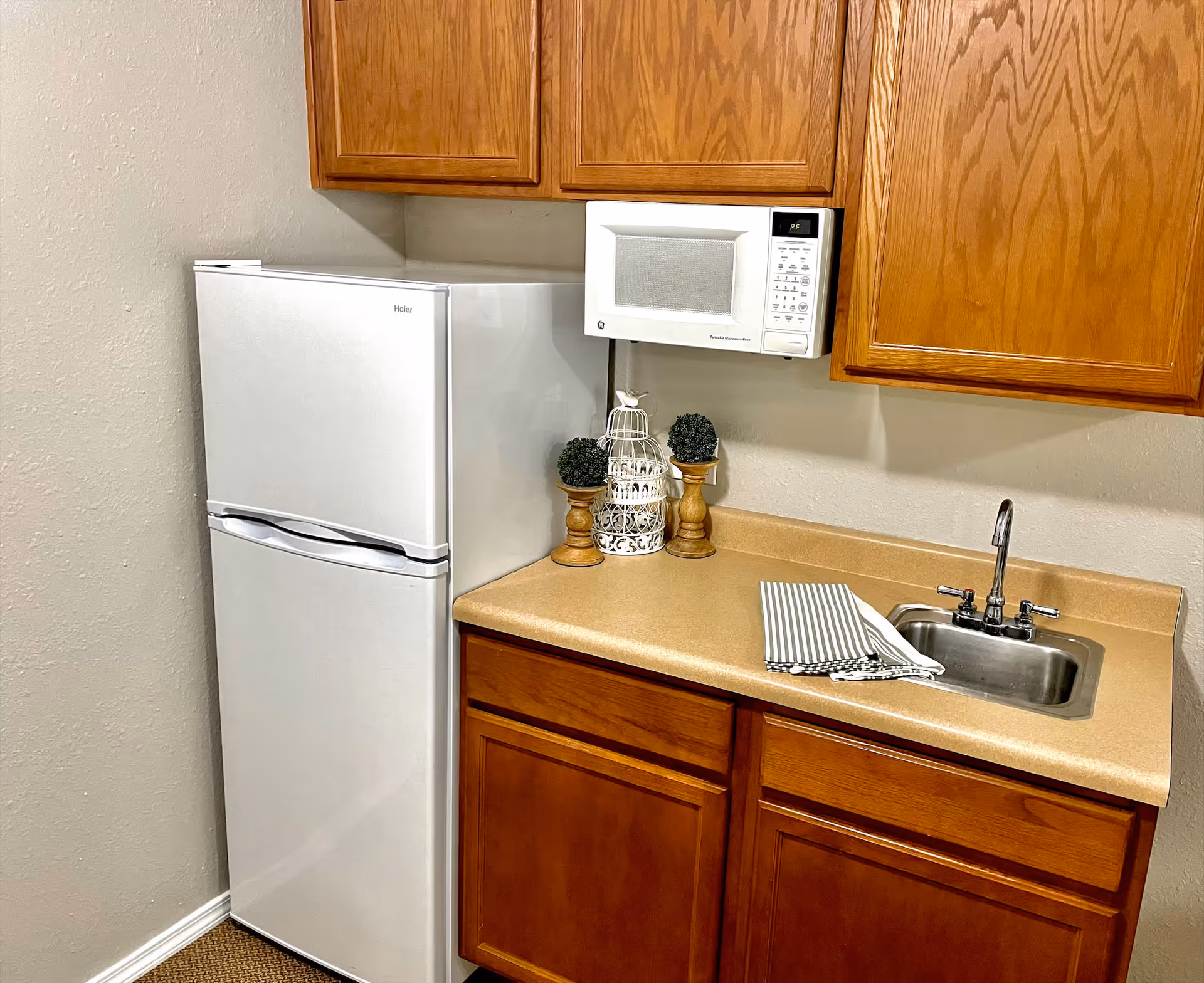A small kitchen area with wooden cabinets, a white refrigerator, a white microwave mounted above the countertop, a stainless steel sink with a faucet, and a striped dish towel on the counter. Decorative items including two small topiary plants and a white birdcage are placed on the countertop.