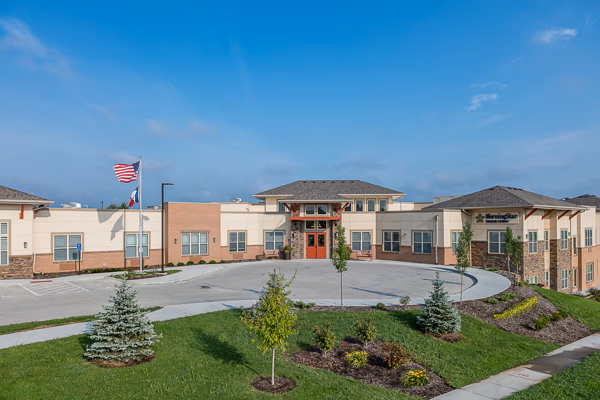 Exterior front of a low-rise assisted living building with a circular driveway, flagpoles, and manicured landscaping under a blue sky.