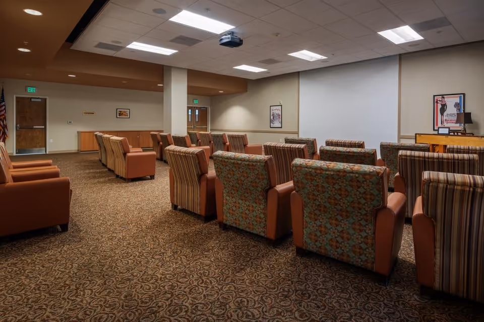 Carpeted common room with rows of upholstered armchairs facing a large projection screen and ceiling projector.