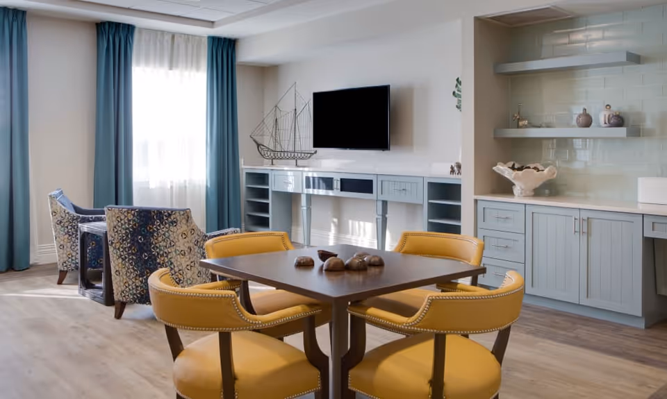 A bright and modern living area with a square wooden table surrounded by four yellow cushioned chairs. In the background, there are two patterned armchairs near a window with blue curtains. A flat-screen TV is mounted on the wall above a blue console table with open shelves. To the right, there is a built-in cabinet with light blue drawers and shelves, decorated with small items and a large shell-shaped bowl.