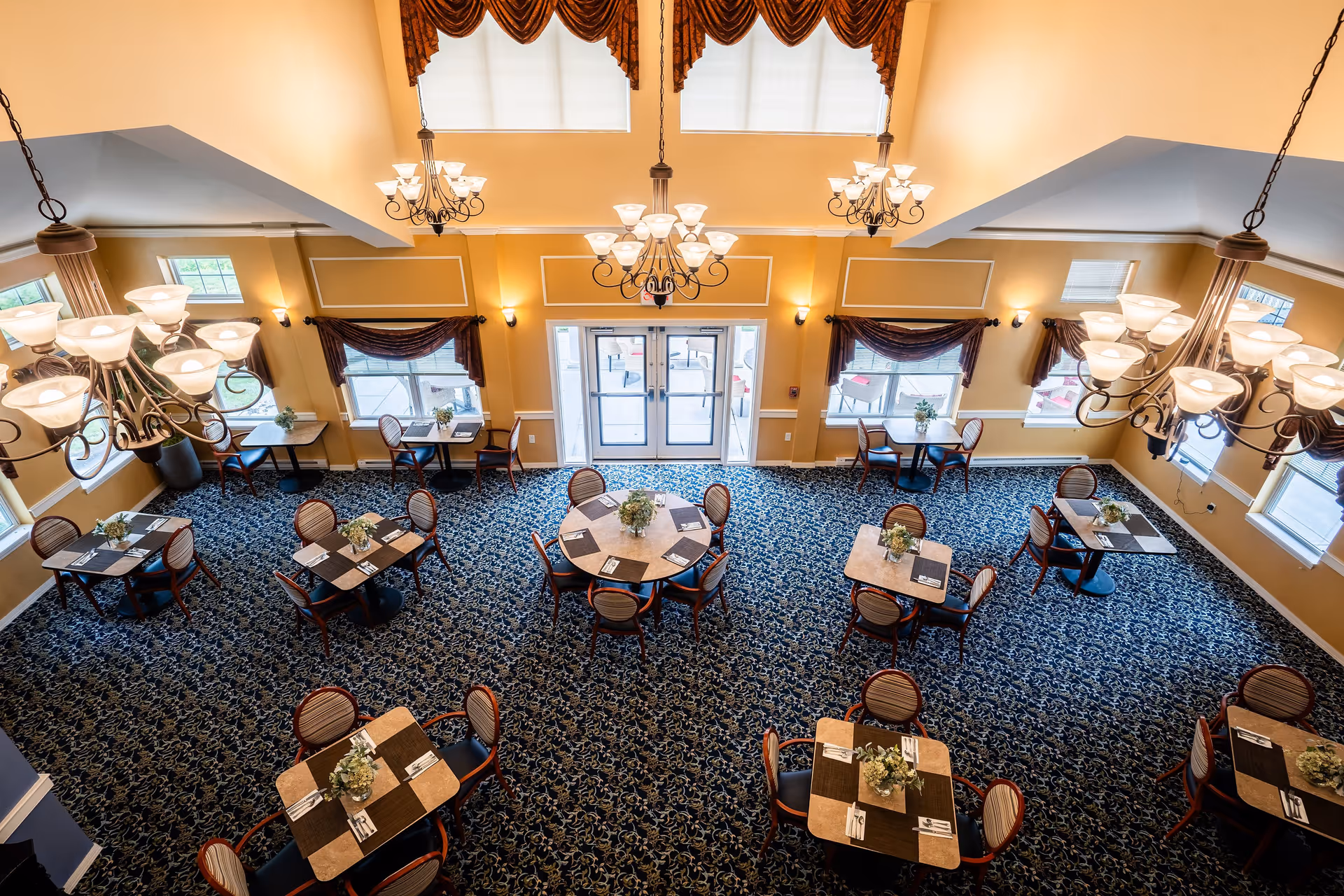 A spacious dining room with multiple tables and chairs arranged neatly on a patterned carpet. The room features large windows with dark curtains, elegant chandeliers hanging from the ceiling, and a set of double glass doors leading outside.