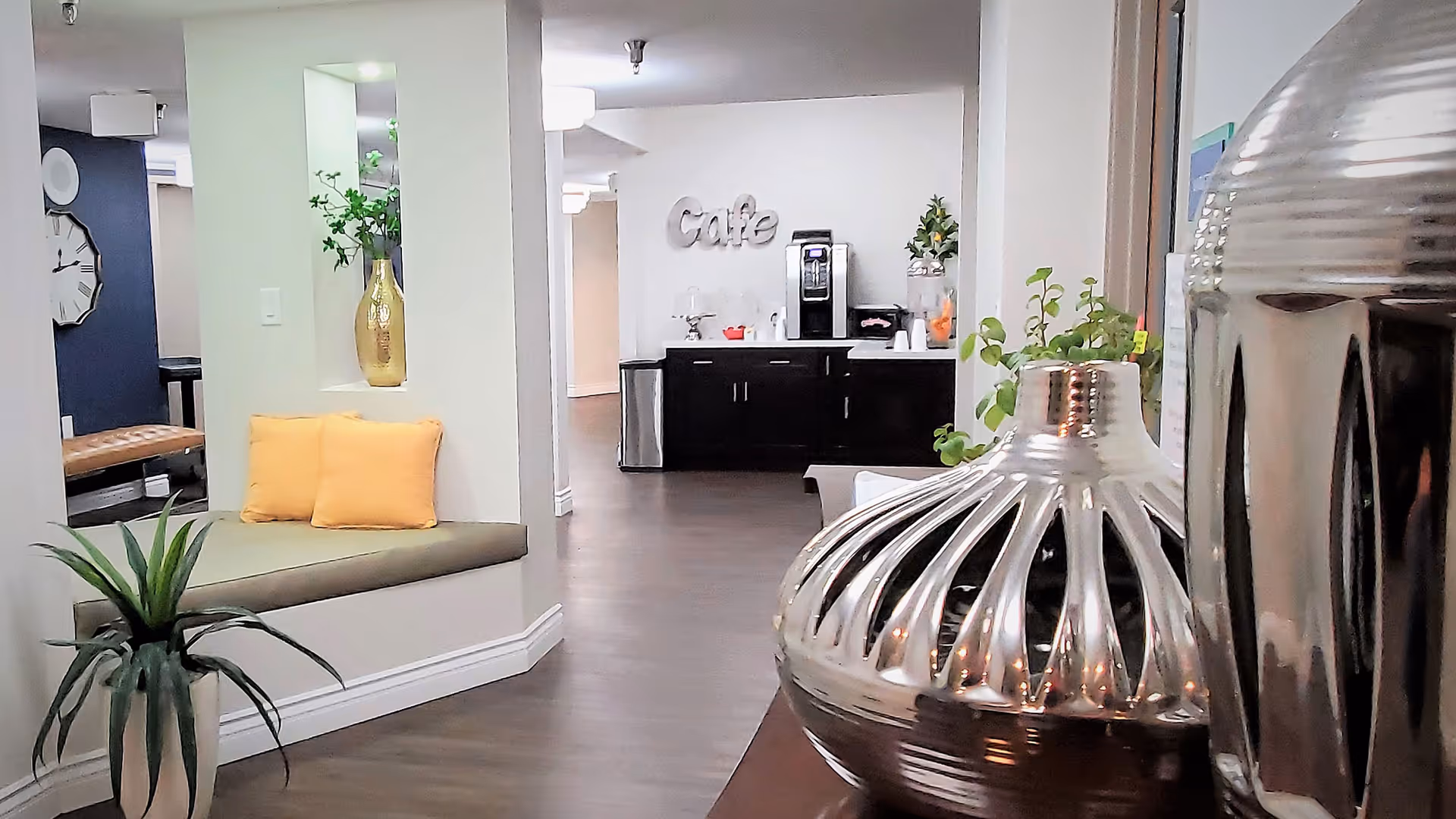Interior view of a senior living facility showing a cozy seating nook with two yellow cushions and a gold vase with greenery. In the background, there is a coffee station with a coffee machine, cups, and a water dispenser. The space has wood flooring and neutral-colored walls with decorative plants and modern decor.