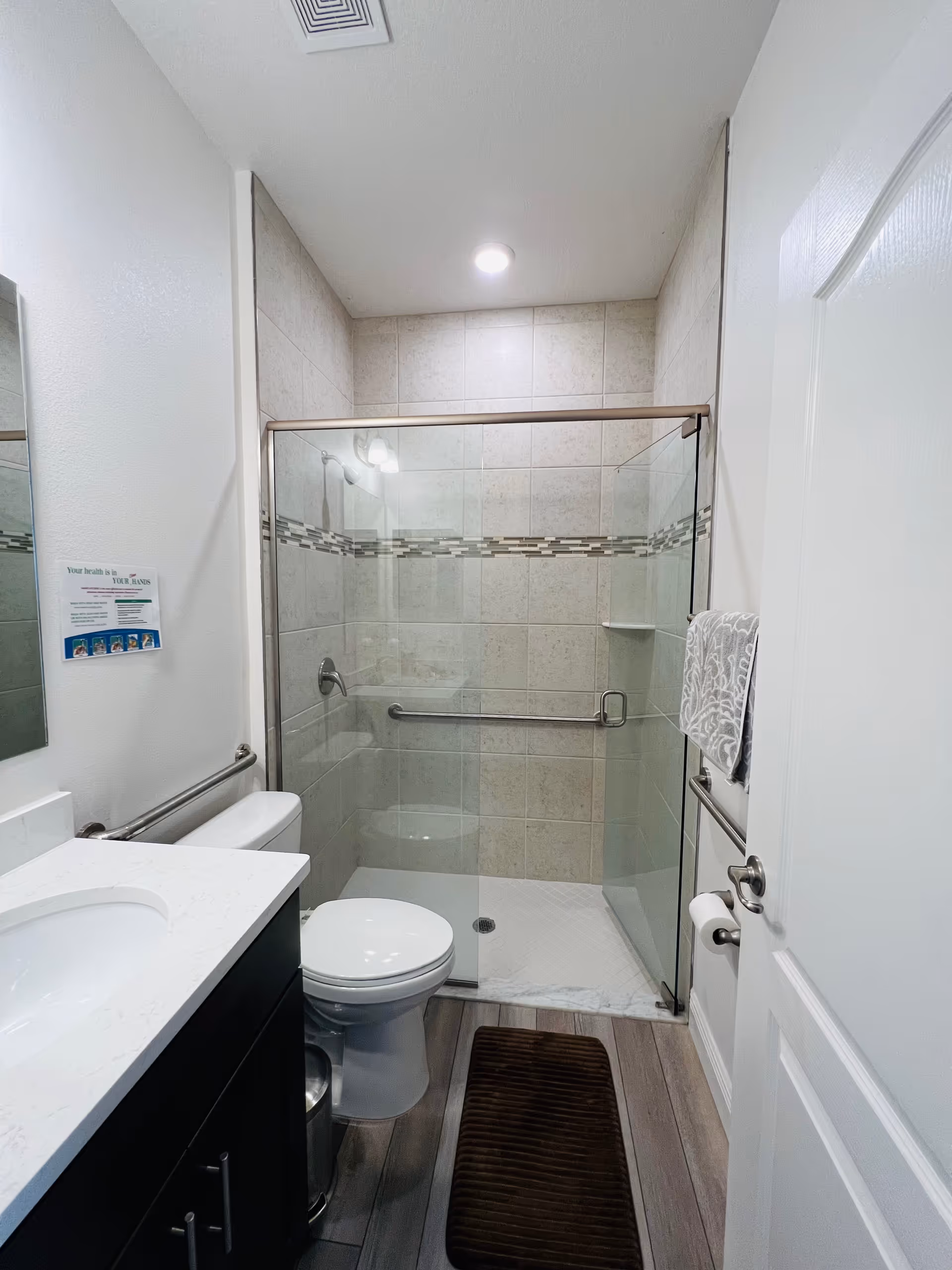 A clean and modern bathroom featuring a glass-enclosed shower with beige tiles and a decorative tile border. The bathroom includes a white toilet, a dark wood vanity with a white countertop and sink, a wall-mounted mirror, a towel rack with a patterned towel, and a brown floor mat on wood-look flooring.