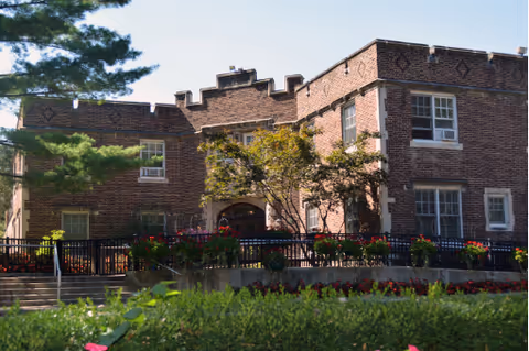 Brick two-story building with a central arched entrance, decorative roofline, and landscaped front with a fenced patio.