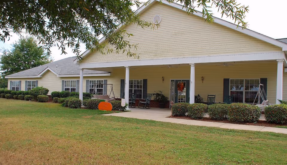 Exterior view of a single-story yellow building with a covered porch featuring rocking chairs and swings. The building is surrounded by neatly trimmed bushes and a grassy lawn, with a tree branch partially visible in the foreground.