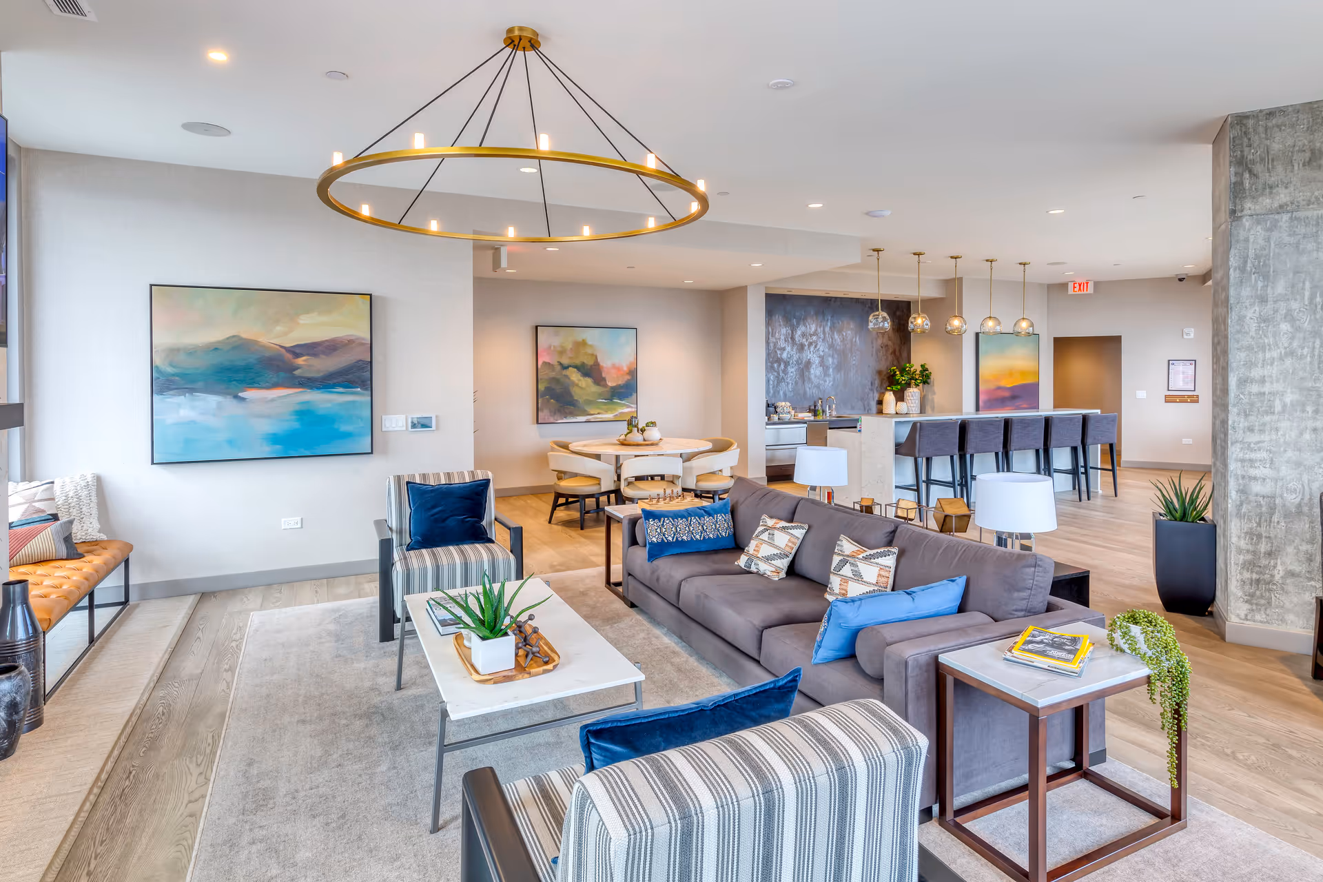 Modern open-concept living area in Avidor Evanston, featuring a gray sofa with decorative pillows, two striped armchairs, a white coffee table with a plant, a round dining table with chairs, and a kitchen island with bar stools and pendant lights. The room has light wood flooring, abstract wall art, and a large circular chandelier.