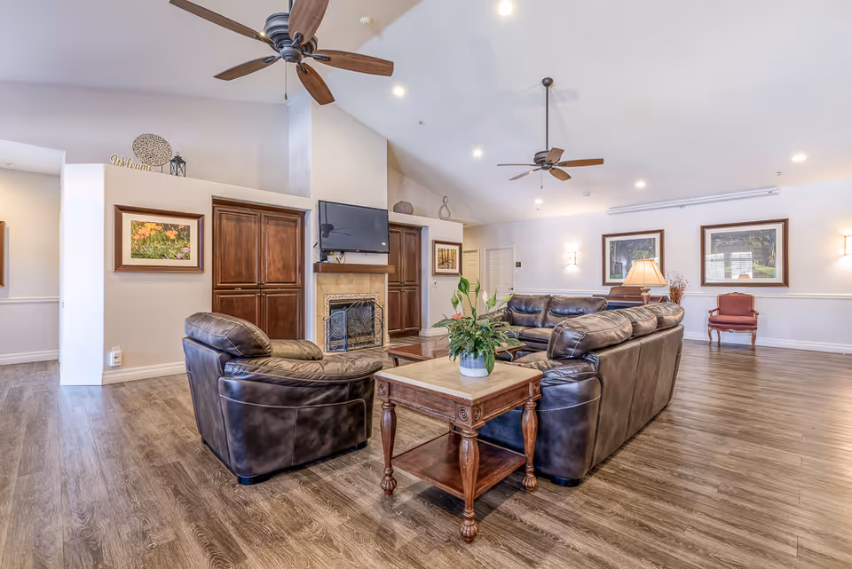 A spacious senior living facility common area with brown leather sofas arranged around a wooden coffee table with a potted plant. The room features a fireplace with a mounted flat-screen TV above it, wooden cabinets on either side, ceiling fans, framed artwork on the walls, and warm lighting fixtures. The floor is covered with wood-style flooring.
