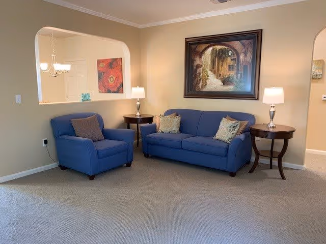 A cozy living room area with a blue armchair and a blue sofa, each with decorative pillows. There are two wooden side tables with lamps on either side of the sofa. A framed painting depicting an archway and pathway hangs on the beige wall behind the sofa. The room has beige carpeting and a pass-through window opening to another room with a chandelier and wall art.