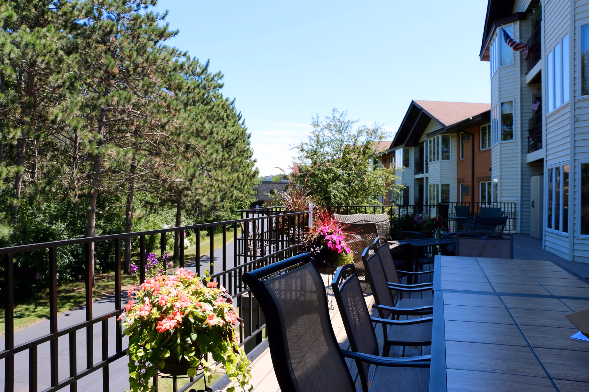 Balcony terrace with a long dining table, chairs, and potted flowers overlooking trees and the building exterior.