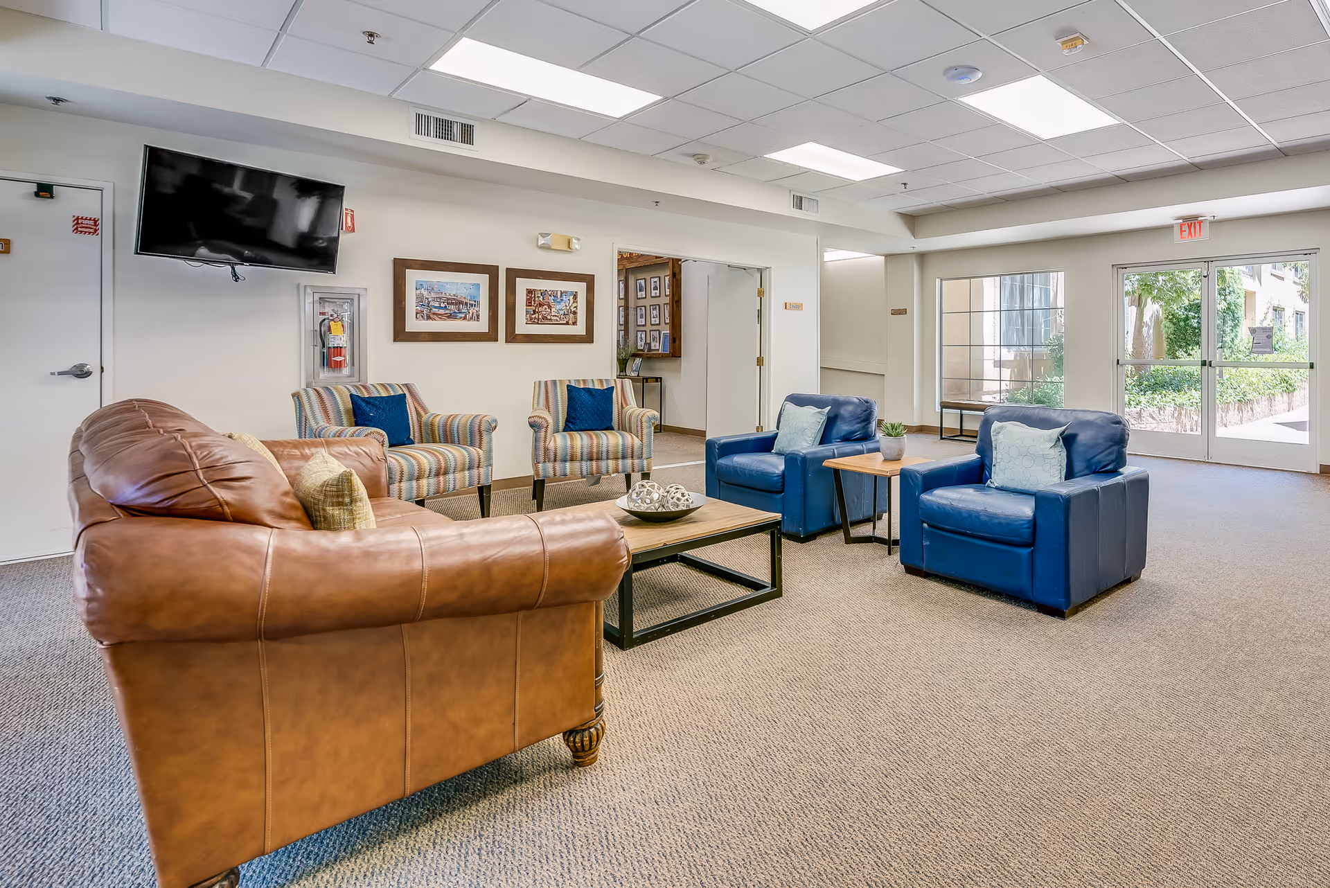 Spacious senior living common room with leather sofa, upholstered and blue armchairs around a coffee table, a wall-mounted TV, and glass exit doors.