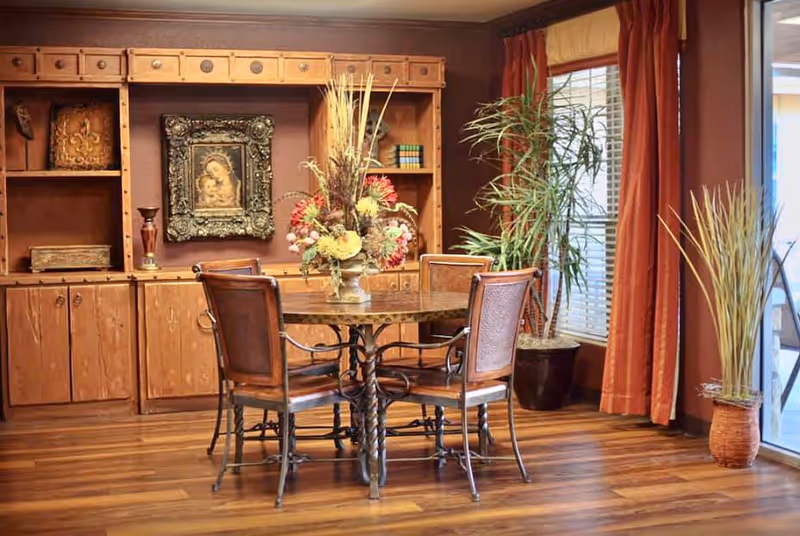 A cozy dining area with a round wooden table surrounded by four chairs with metal frames and cushioned seats. The table has a large floral centerpiece. Behind the table is a wooden built-in cabinet with shelves, some decorative items, and a framed picture. To the right, there is a large window with orange curtains and two potted plants on the floor. The room has wooden flooring and warm lighting.