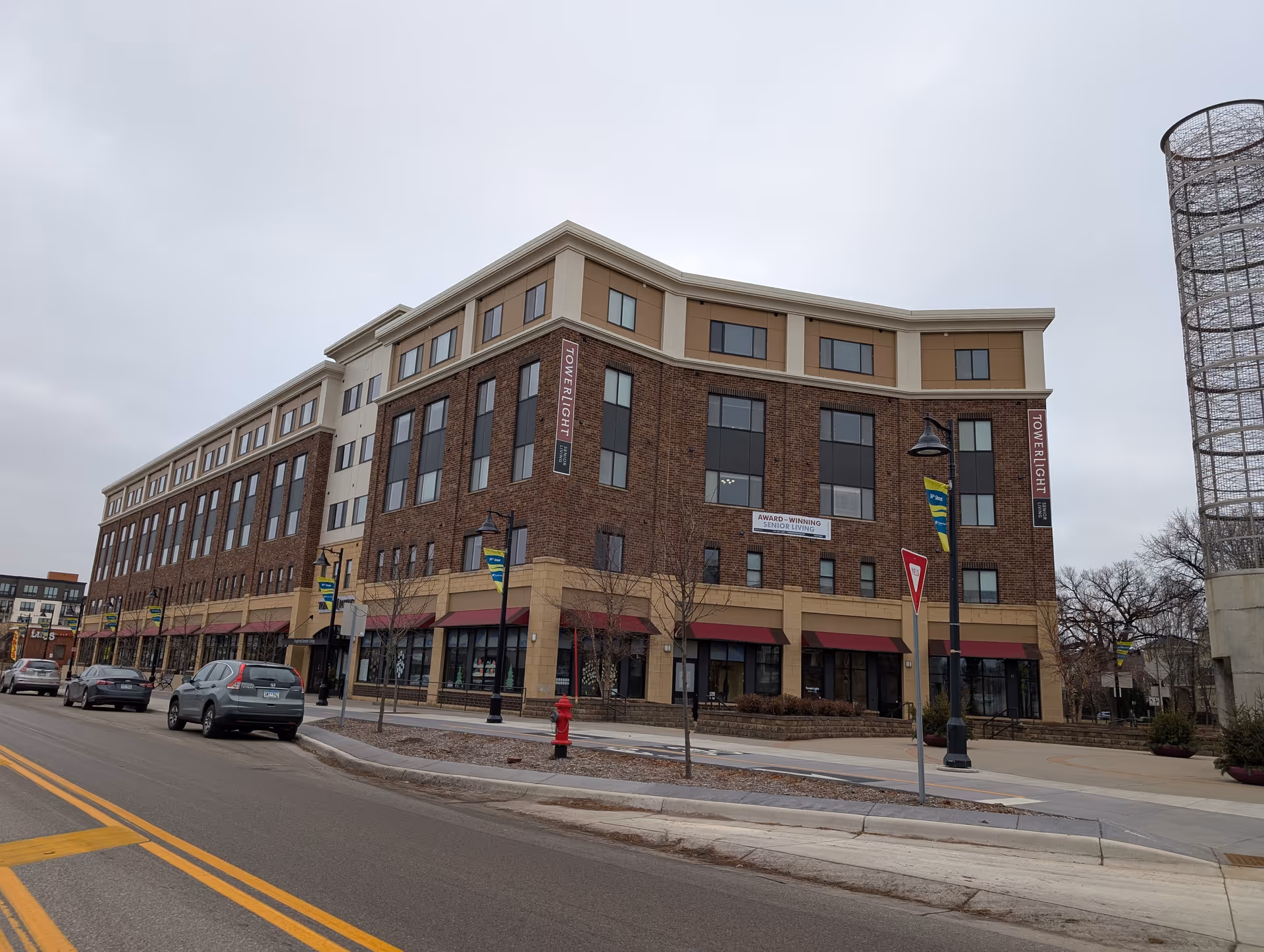 Exterior view of a multi-story brick and beige building with large windows and maroon awnings, located on a street corner with parked cars and street lamps. The building has signs reading 'TOWERLIGHT' and 'AWARD-WINNING SENIOR LIVING'.