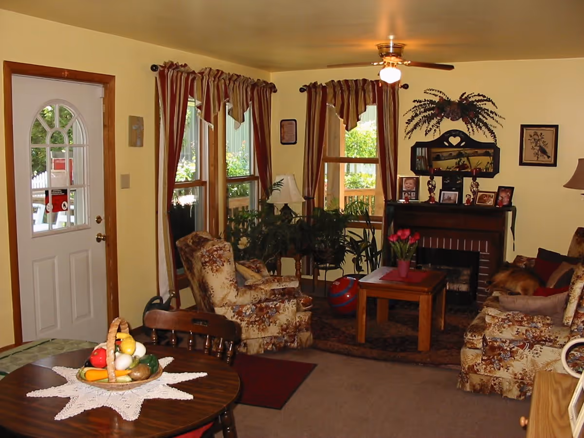 A cozy living room with floral patterned armchairs and sofa, a wooden coffee table with a vase of red tulips, a fireplace decorated with framed photos and a wall-mounted plant above it. The room has yellow walls, three windows with red and beige striped curtains, a ceiling fan with a light, and a round wooden dining table with a basket of artificial fruits on a white doily. A white door with a window is visible on the left side.