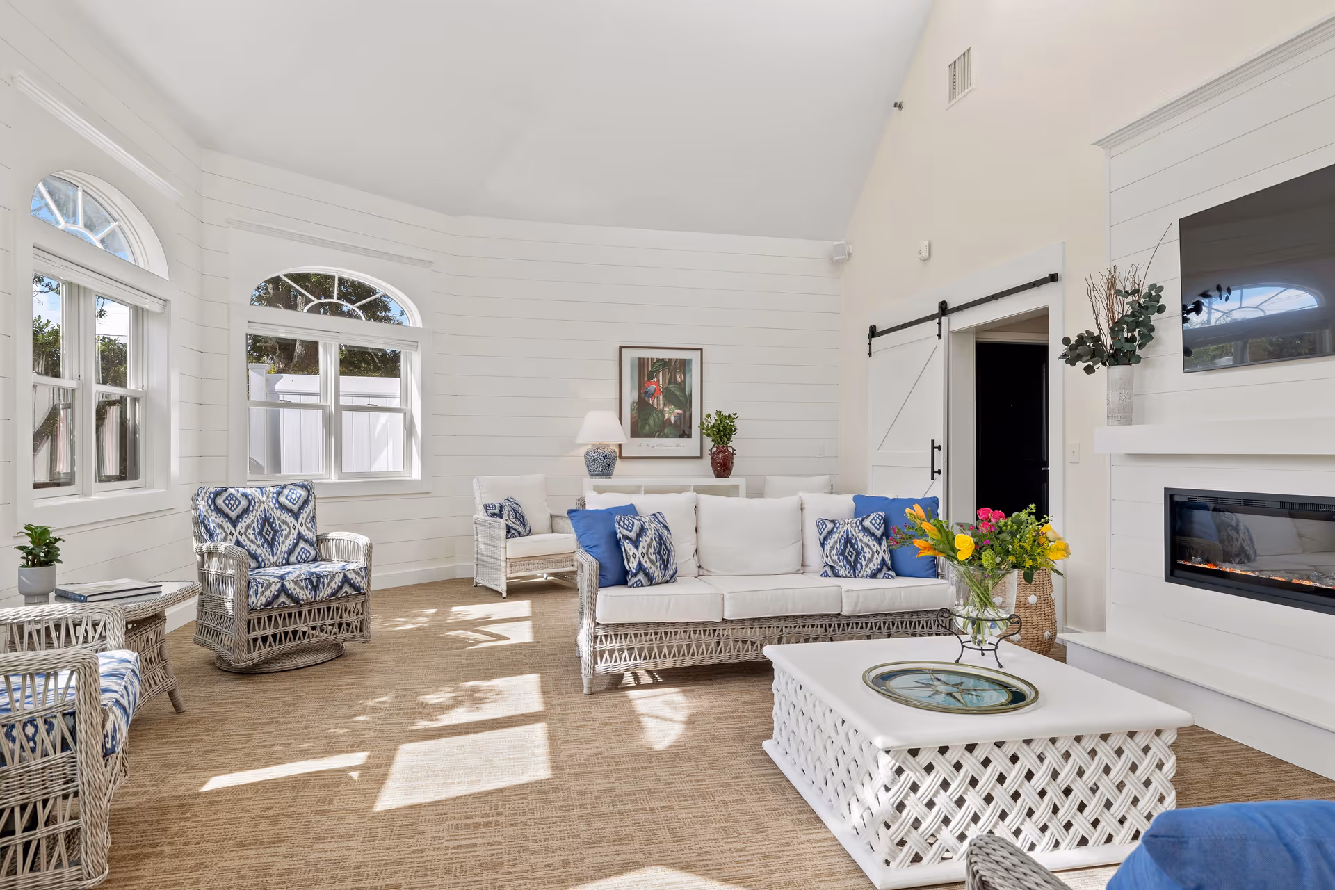 Bright and airy living room with white wicker furniture including a sofa and chairs with blue and white cushions. The room features large windows with arched tops letting in natural light, a white coffee table with a decorative tray, a fireplace with a mounted TV above it, and a sliding barn door. The walls are white with horizontal paneling and there are decorative plants and a framed picture on the wall.