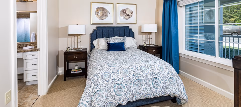 Bright furnished bedroom with a patterned blue-and-white bed, dark nightstands and lamps, a window with blue curtains, and an open door to a bathroom.
