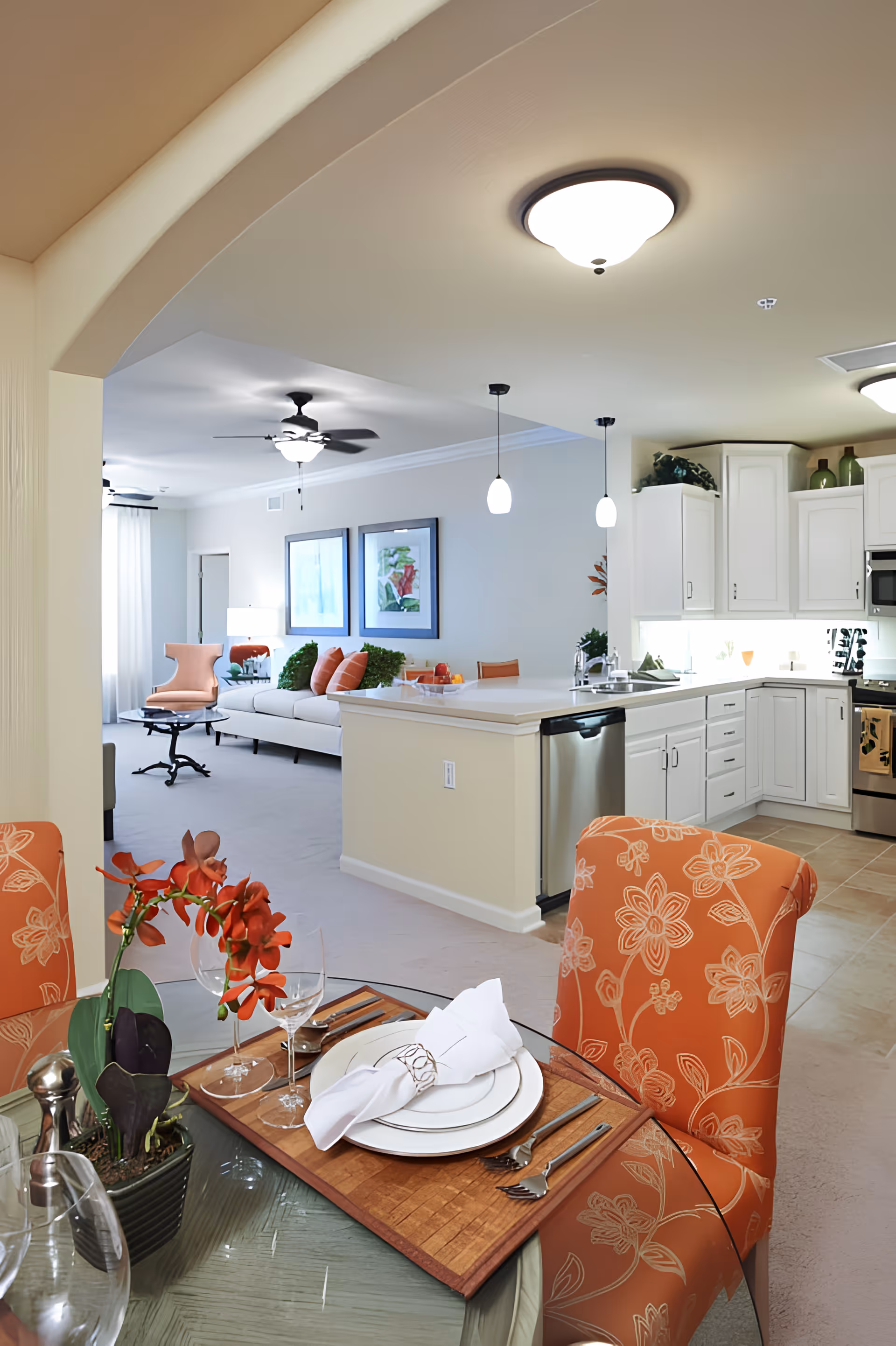 Open-plan interior with a set dining table and orange patterned chairs in the foreground, a kitchen island and white cabinets to the right, and a living area with sofa and chairs in the background.