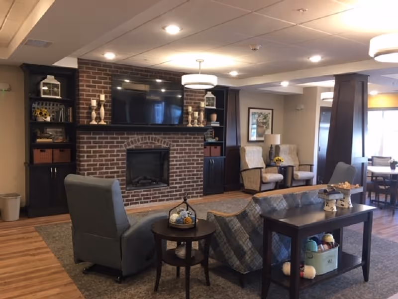 A cozy living room area featuring a brick fireplace with a mounted flat-screen TV above it. The fireplace is flanked by built-in dark wood shelves and cabinets decorated with various items including candles and lanterns. In front of the fireplace, there are comfortable upholstered chairs and a sofa arranged around a small round table and a rectangular console table with decorative items and yarn. The room has warm lighting and wood flooring, with additional seating and a dining area visible in the background.