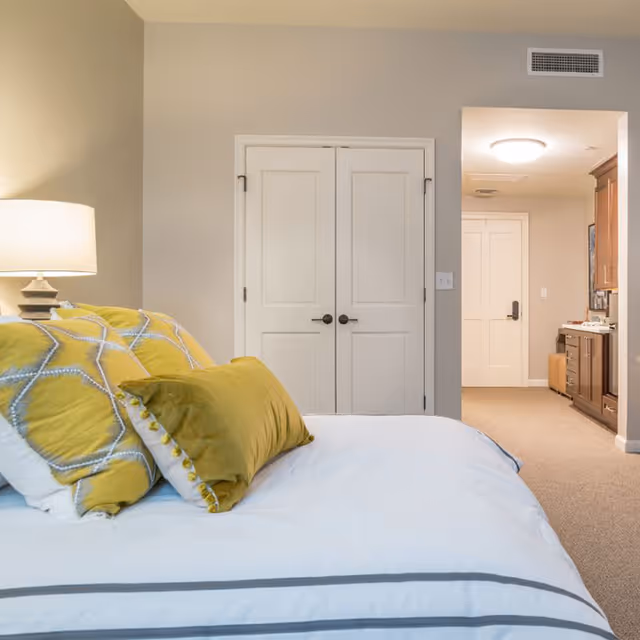 A bedroom with a neatly made bed featuring white and yellow pillows. A bedside lamp is on the left side. There is a double-door closet in the center of the wall and an open doorway leading to a hallway and kitchen area with wooden cabinets on the right.
