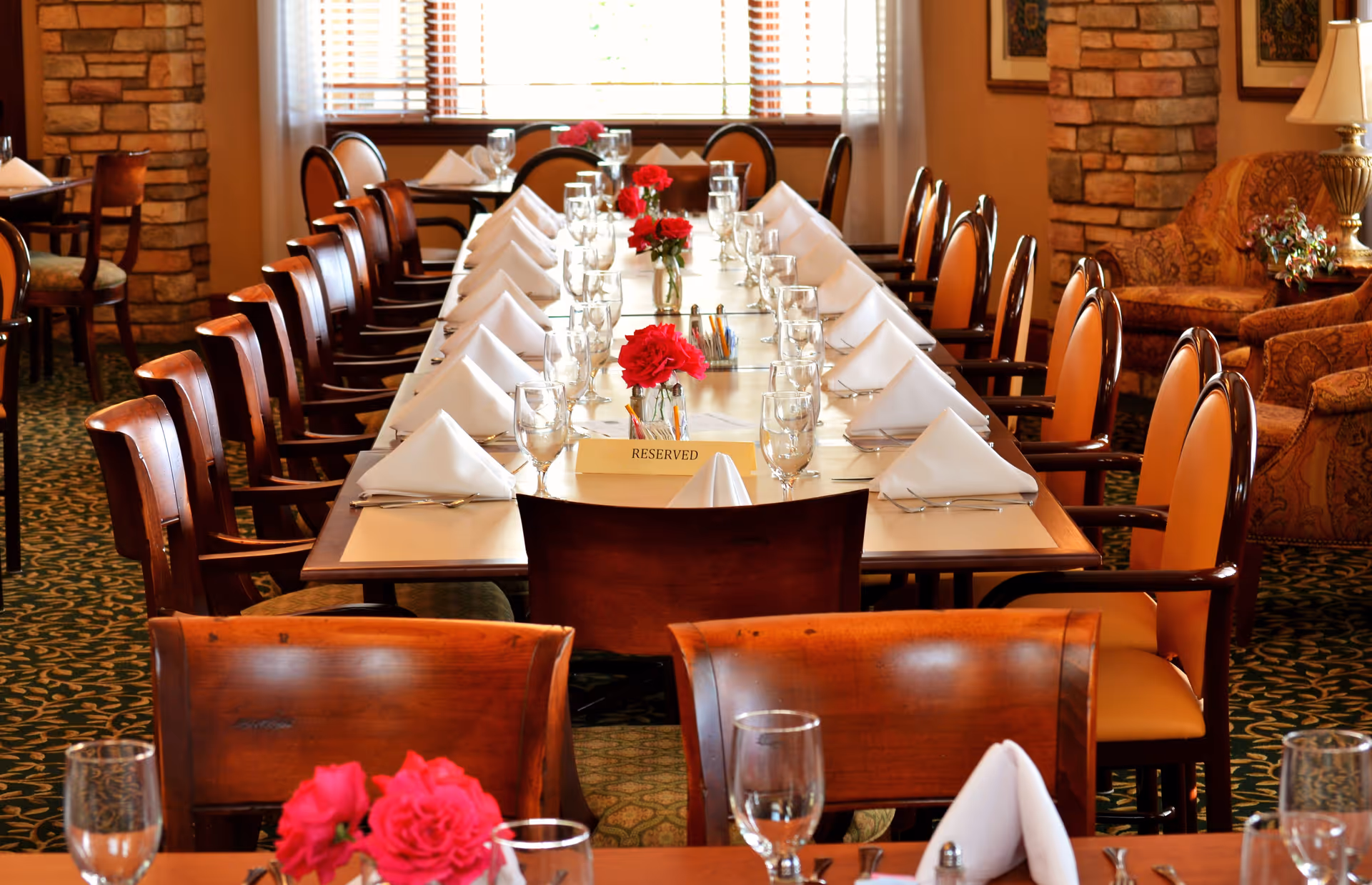 Long dining table set for a meal with folded napkins, glassware, red flower centerpieces and a "Reserved" sign in a warm dining room.