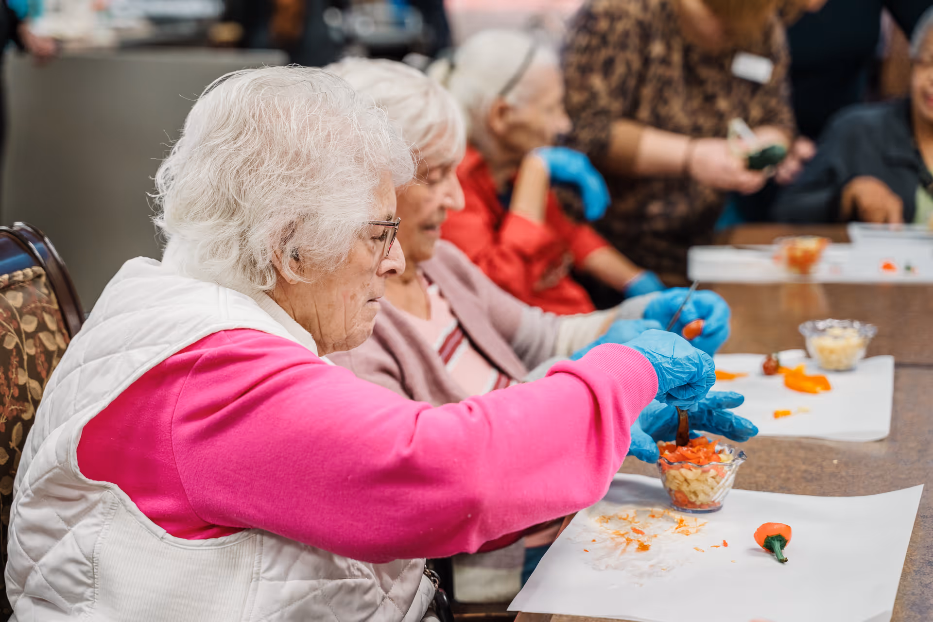 Elderly women sitting at a table engaged in a food preparation activity, wearing blue gloves and placing chopped vegetables into small glass bowls.