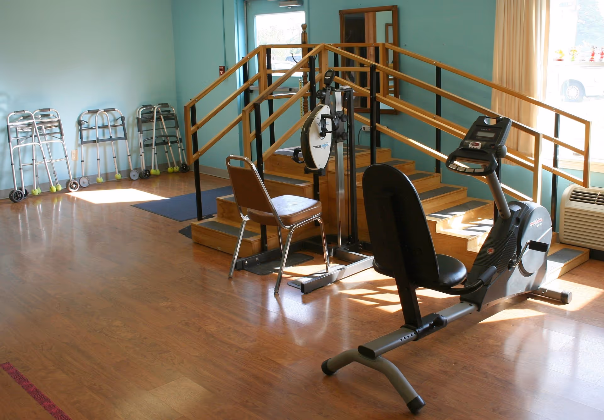 A rehabilitation/exercise room with a recumbent exercise bike, therapy stairs with handrails, and several walkers lined up against a wall.