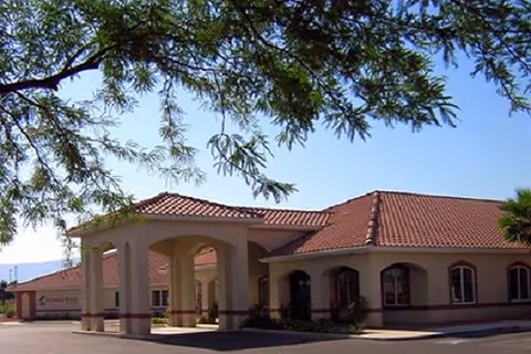Exterior view of Highland Manor of Mesquite, a single-story building with a red-tiled roof and beige walls, featuring an arched entrance and surrounded by a paved driveway and some trees.