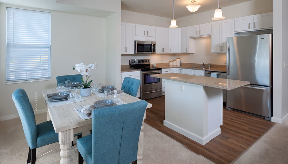 Bright open kitchen with a center island and stainless steel appliances and an adjoining dining table set with teal chairs.