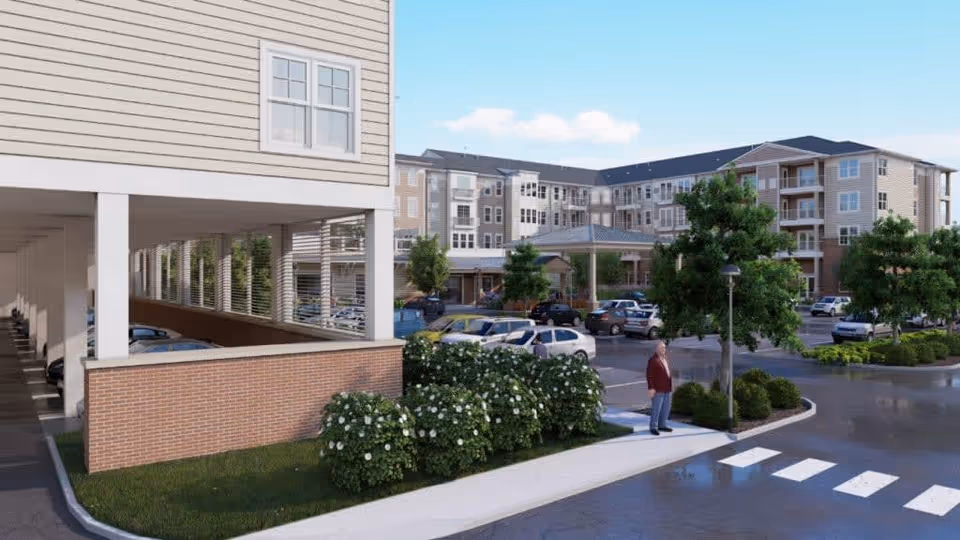 View of the exterior of a senior living facility named Acclaim At East Beach, showing a multi-story building with balconies, a covered parking area, landscaped bushes and trees, a crosswalk, and a person standing near a lamppost in the parking lot.