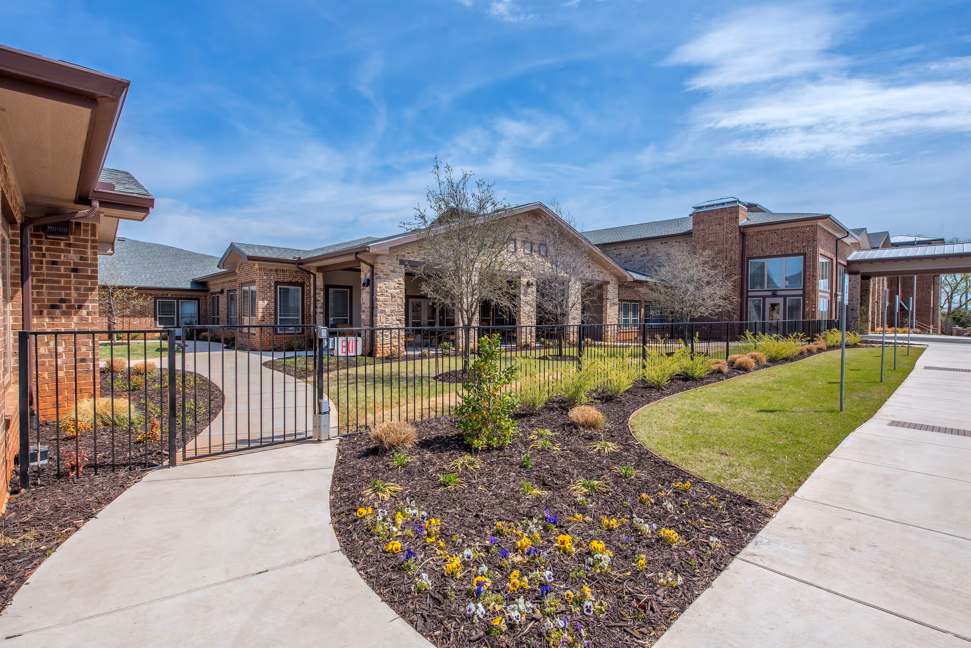 Exterior view of the Legend of Edmond senior living facility showing a brick building with a covered entrance, a black metal fence, landscaped garden beds with flowers and shrubs, and a concrete walkway under a blue sky with some clouds.