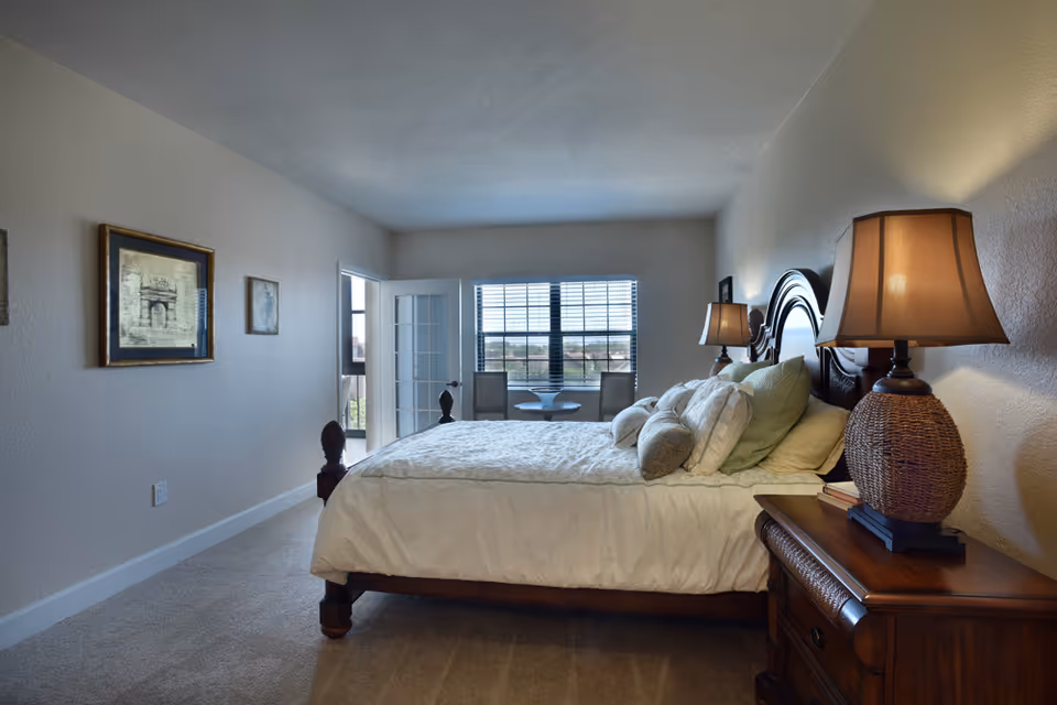 A bedroom with a large bed covered in white bedding and multiple pillows. There are two bedside tables with lamps on each side of the bed. The room has beige walls with framed artwork and a window with blinds letting in natural light. A small table with two chairs is visible near the window.
