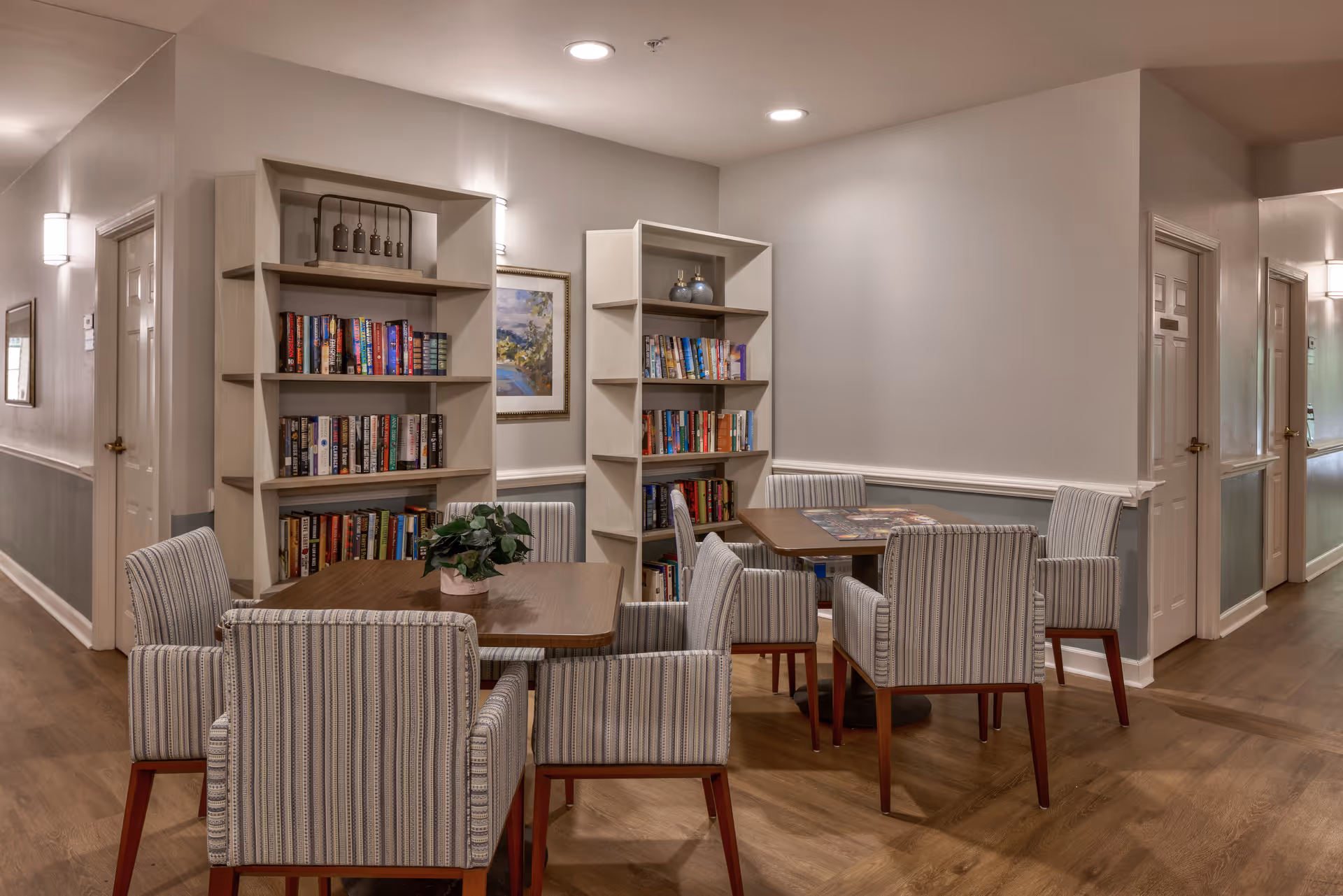 A cozy interior common area with two wooden tables surrounded by striped upholstered chairs. Behind the tables are two bookshelves filled with books and decorative items. The walls are painted in neutral tones with framed artwork and soft lighting fixtures. The floor is wooden, and there are doors and a hallway visible in the background.