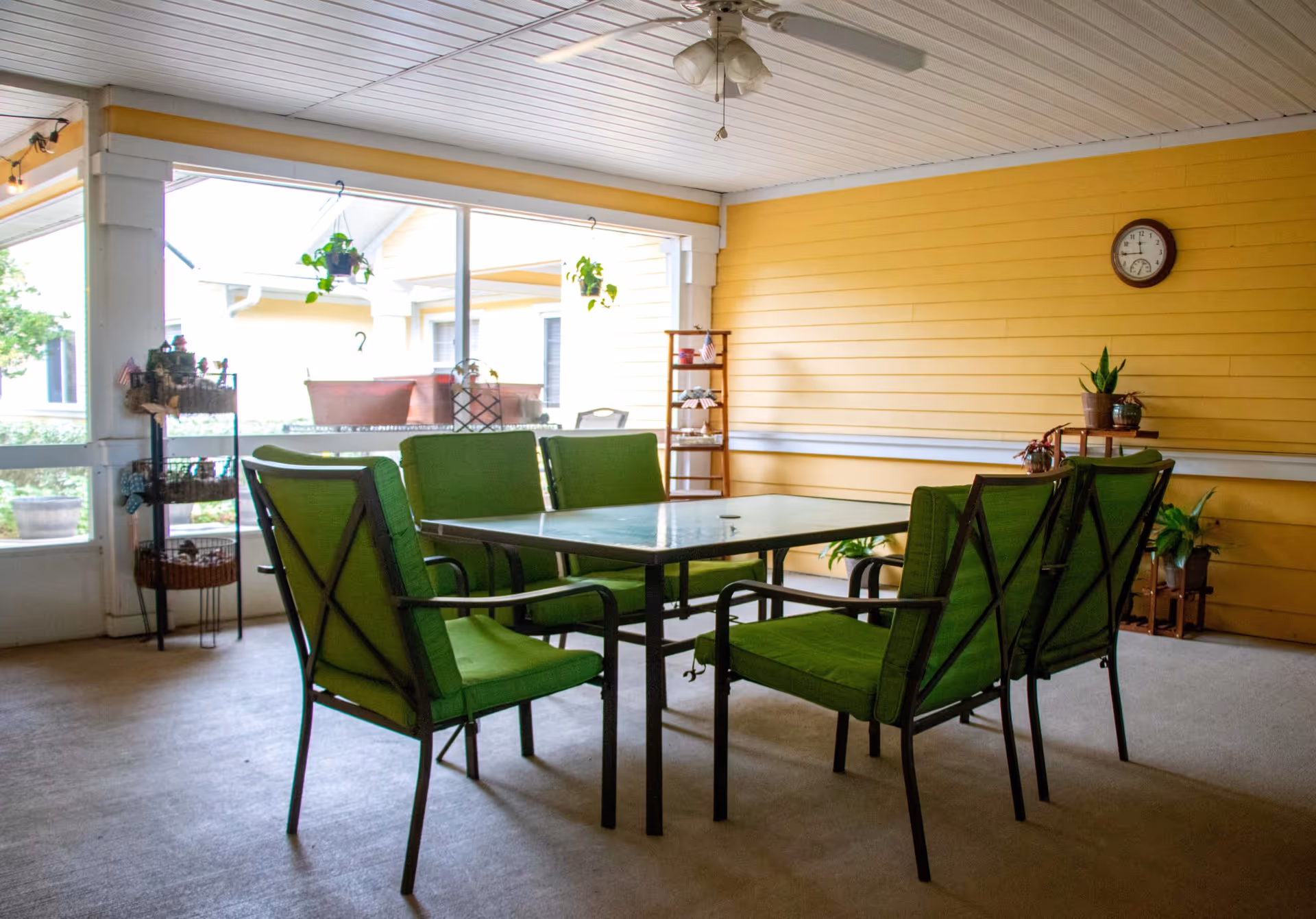 Enclosed sunroom-style common area with a glass-top table surrounded by six green cushioned chairs, yellow paneled wall, and several potted plants.