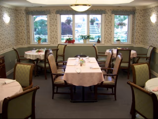A cozy dining room with several tables covered in light pink tablecloths, each set with napkins and utensils. The room has patterned wallpaper, green wainscoting, and large windows with a view of greenery outside. Wooden chairs with cushioned seats surround the tables.