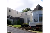 Exterior view of a light-colored building with a sloped driveway and a sign in front that reads 'Envoy of Staunton'. The building has large windows and a small landscaped area with bushes and flowers.