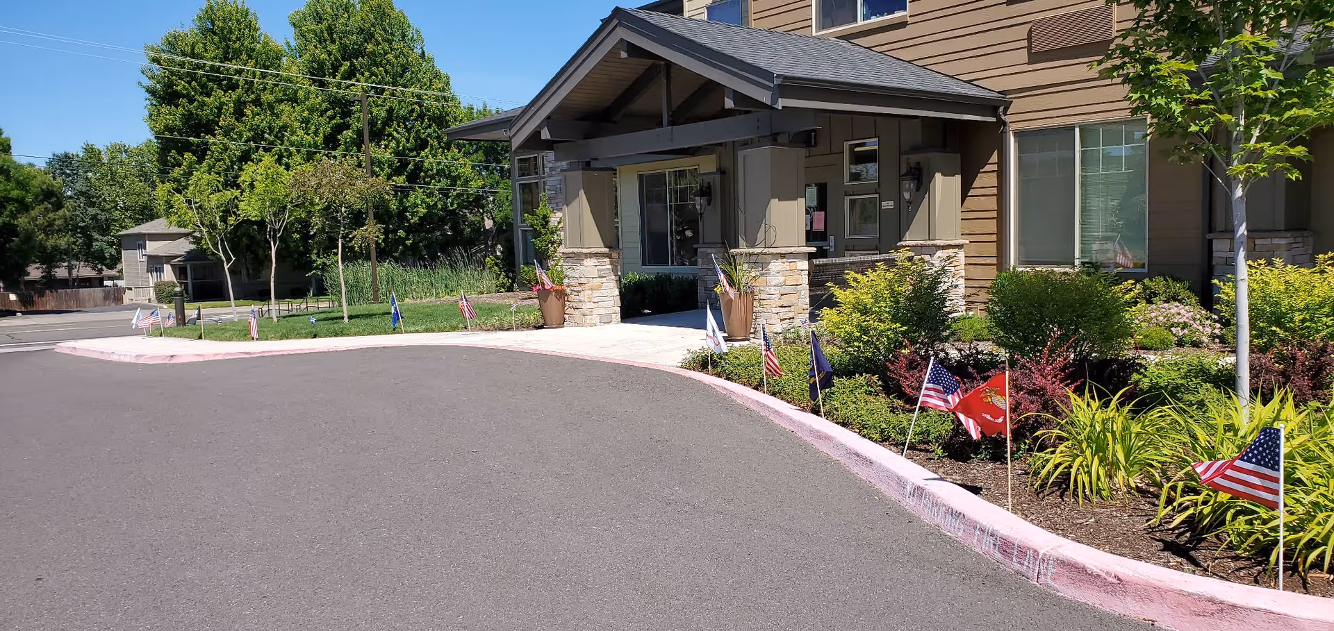 Covered main entrance and driveway of a senior living building with landscaped beds and small American flags along the curb.