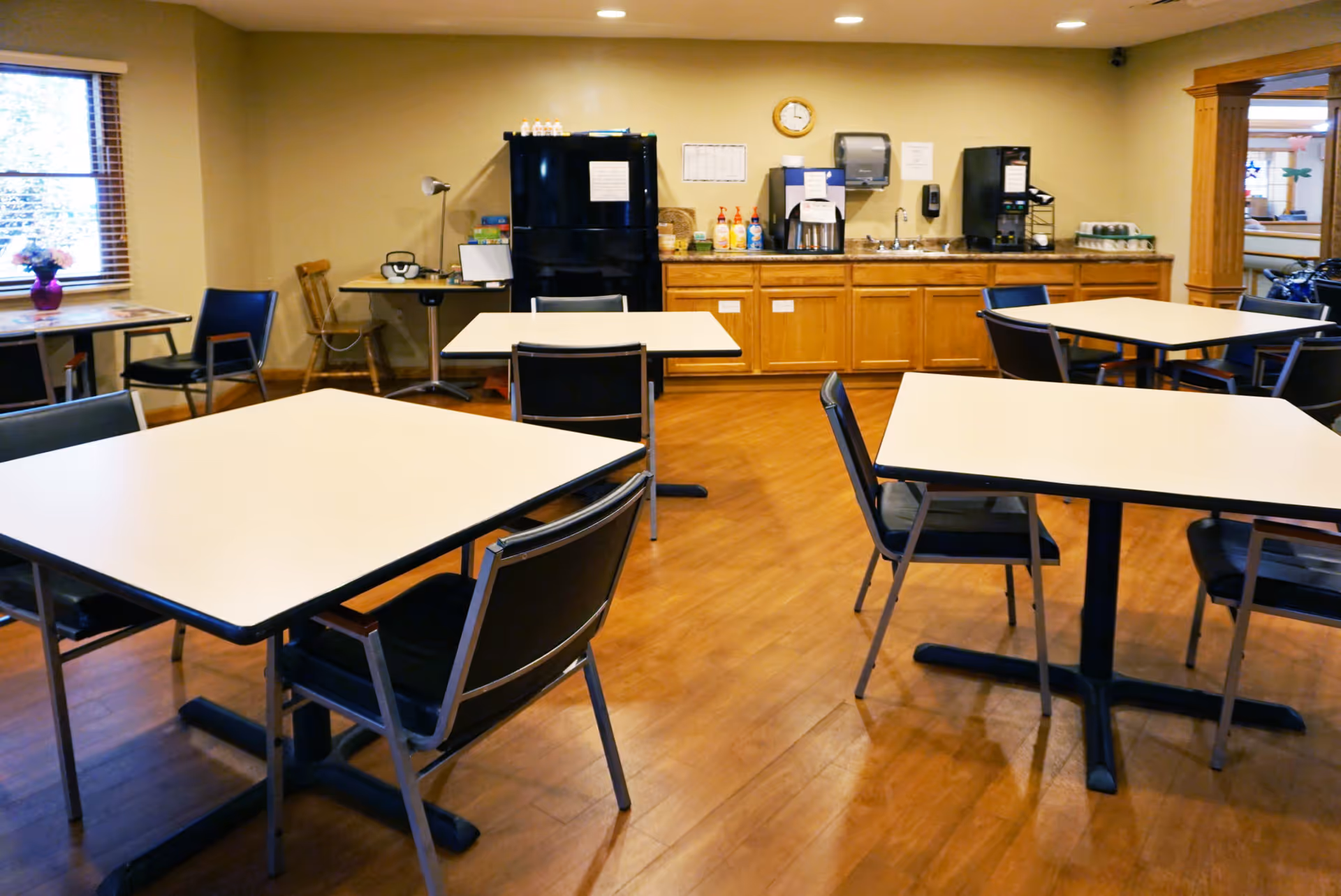 Interior view of a dining area in Snyder Village Assisted Living with several square tables and black chairs arranged on a wooden floor. In the background, there is a counter with a black refrigerator, coffee machines, a sink, and various supplies. A window with blinds and a small table with a vase of flowers are visible on the left side.