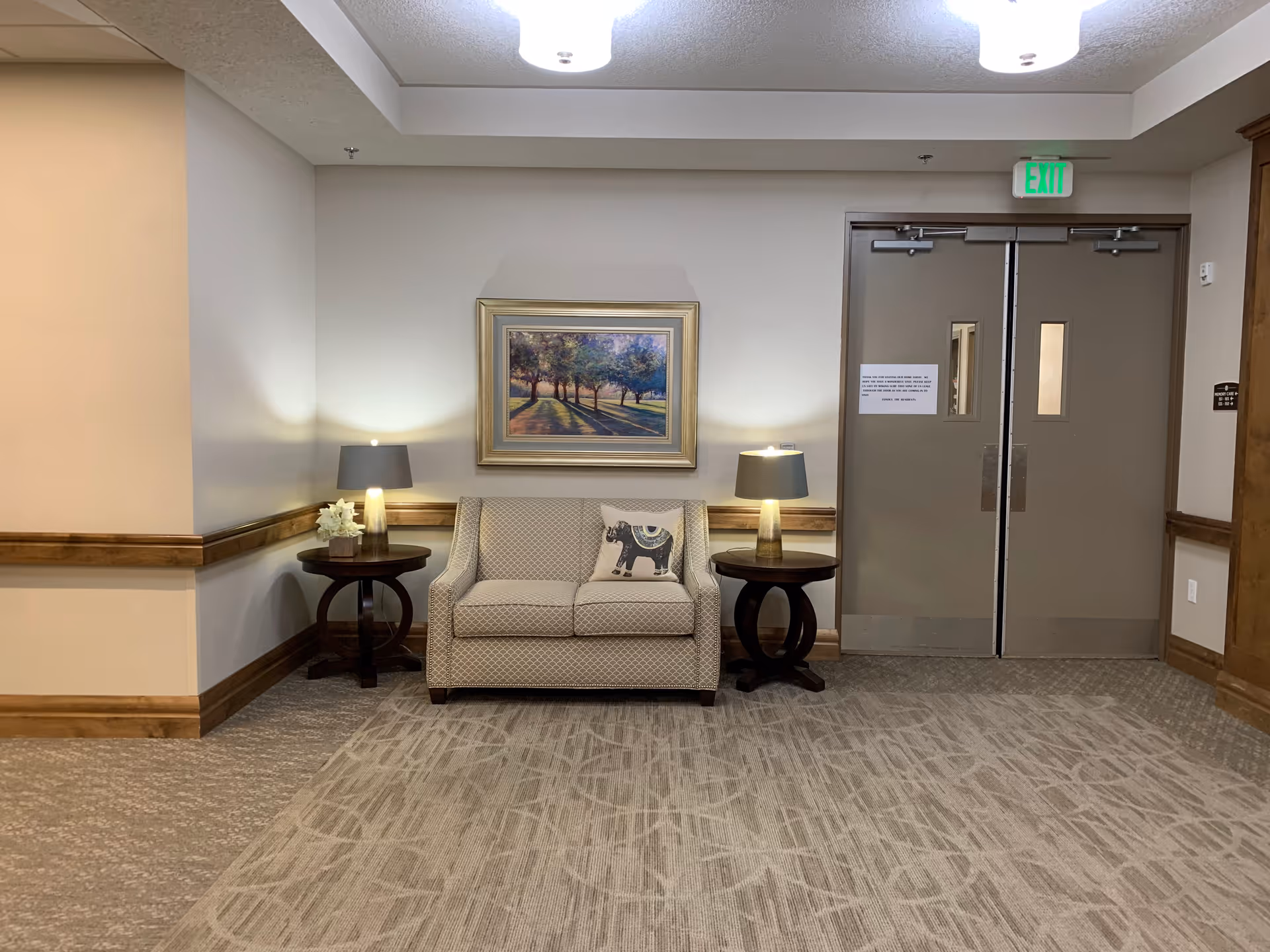 A small seating area in a hallway with a beige loveseat featuring a decorative pillow, flanked by two round wooden side tables each with a lamp and a small flower arrangement. Above the loveseat is a framed painting of a tree-lined path. To the right are double doors with small windows and an exit sign above.