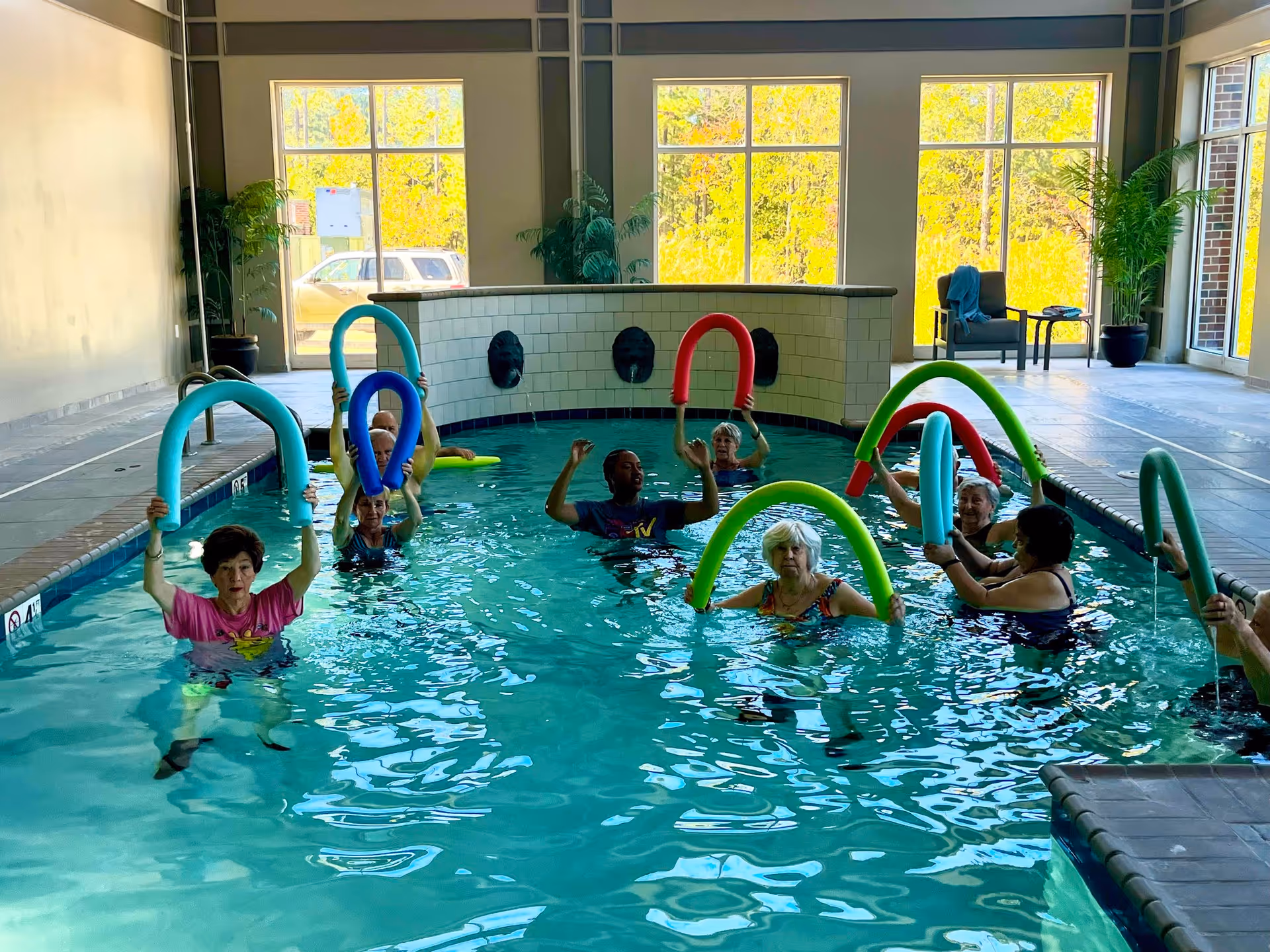 A group of elderly people participating in a water exercise class in an indoor swimming pool, each holding colorful pool noodles above their heads. The pool area has large windows letting in natural light and some potted plants near the walls.