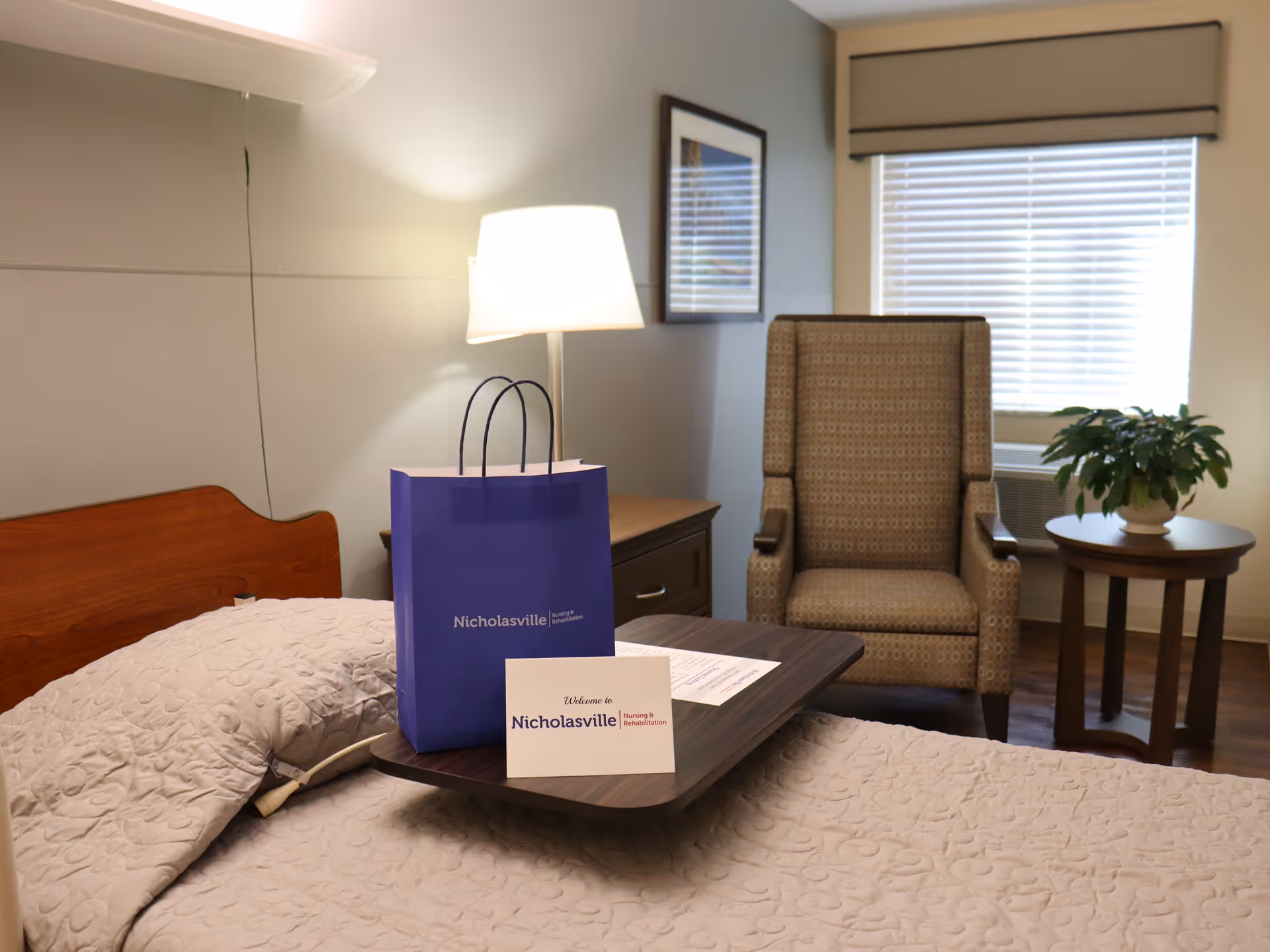 A cozy nursing facility room with a bed covered in a beige quilt, a wooden headboard, and a bedside table with a lamp. On the bed is a wooden tray holding a blue gift bag and a welcome card from Nicholasville Nursing and Rehabilitation. In the background, there is a patterned armchair, a small round table with a potted plant, and a window with blinds and a beige valance.