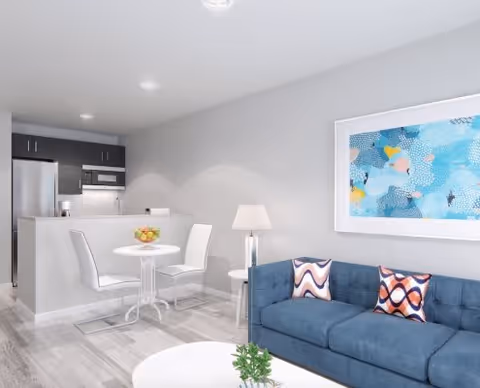 A modern living area with a blue sofa adorned with patterned pillows, a white round coffee table with a small plant, a white round dining table with two white chairs, and a kitchen area with dark cabinets, a refrigerator, microwave, and a bowl of fruit on the dining table. A colorful abstract painting hangs on the wall above the sofa.