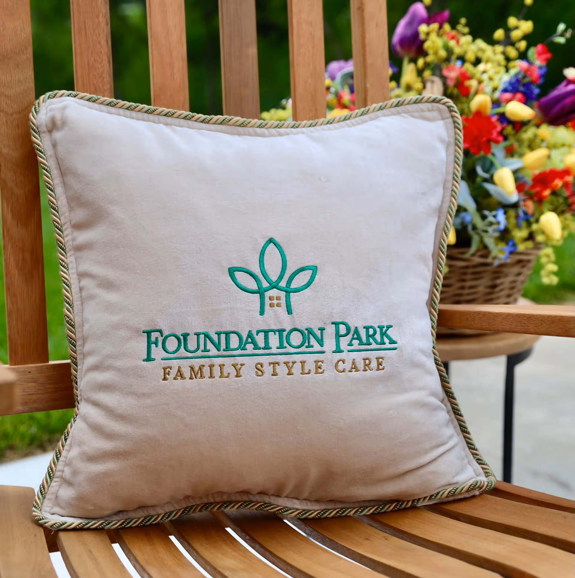 A beige embroidered pillow that reads "Foundation Park Family Style Care" rests on a wooden bench with a basket of colorful flowers in the background.